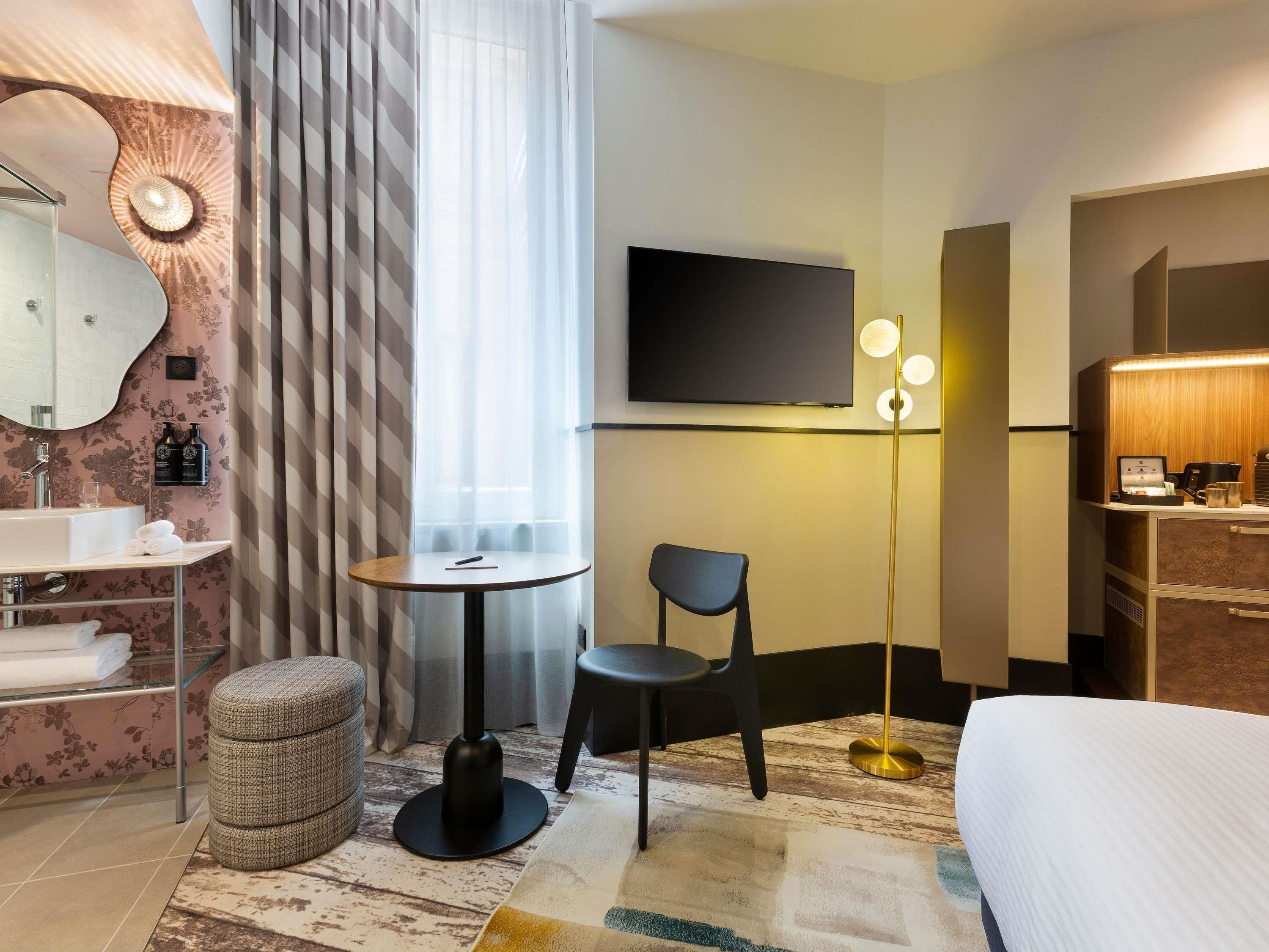 Habitación de hotel moderna con cama, una mesa redonda con silla, televisor, minibar y baño con lavamanos y espejo.