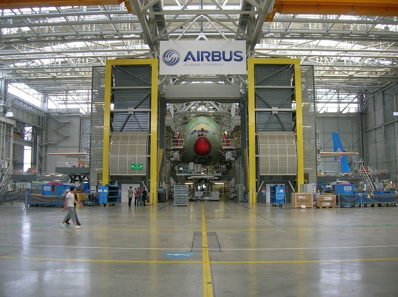 A large industrial hangar with a prominent Airbus logo, a large aircraft in the center, and various equipment and personnel around.