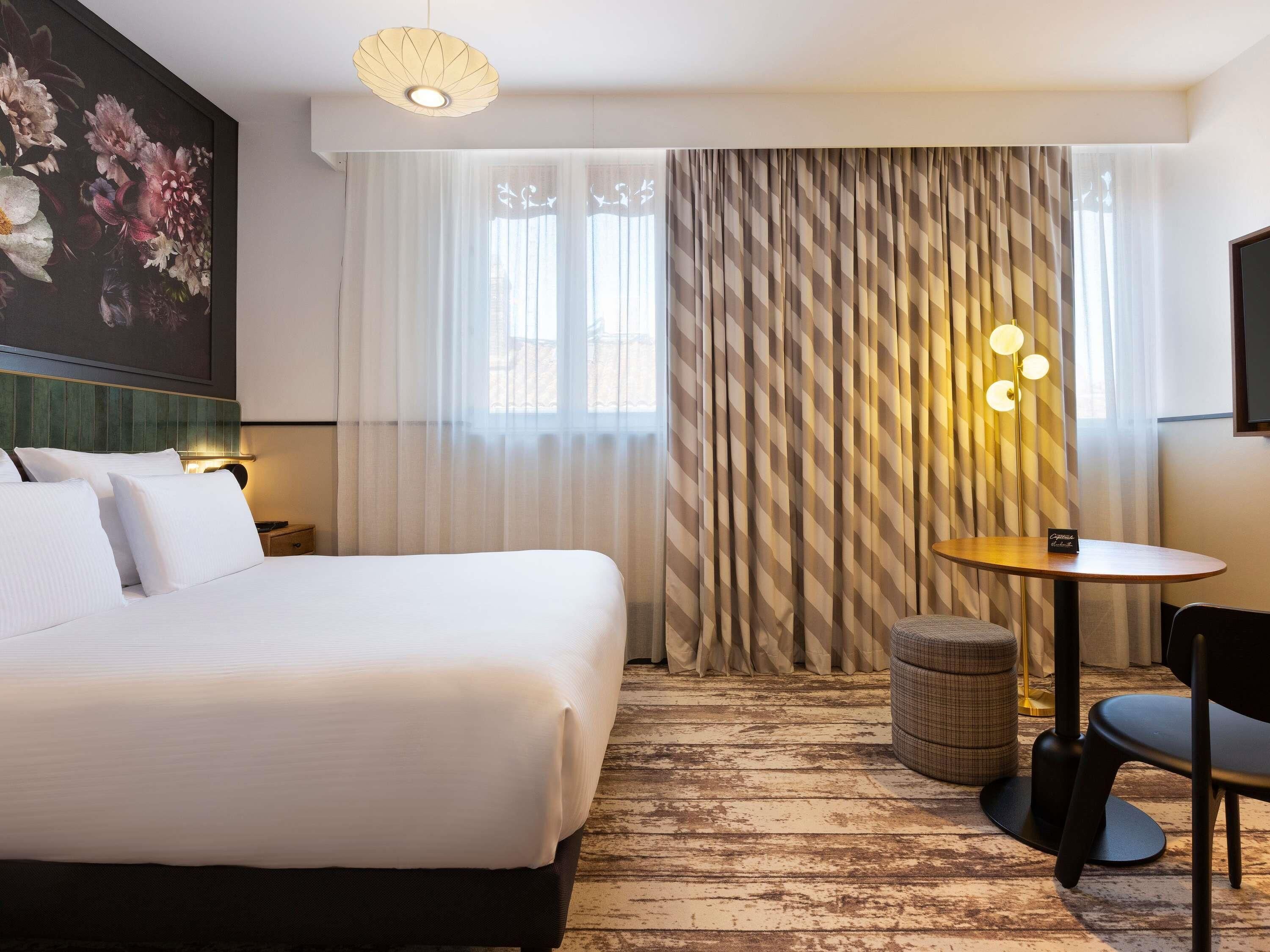 Una habitación de hotel con una cama grande, una mesita, una silla, una lámpara y un cuadro en la pared.