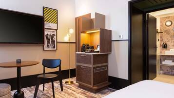Habitación de hotel con cama, escritorio, silla, minibar y televisor en la pared.