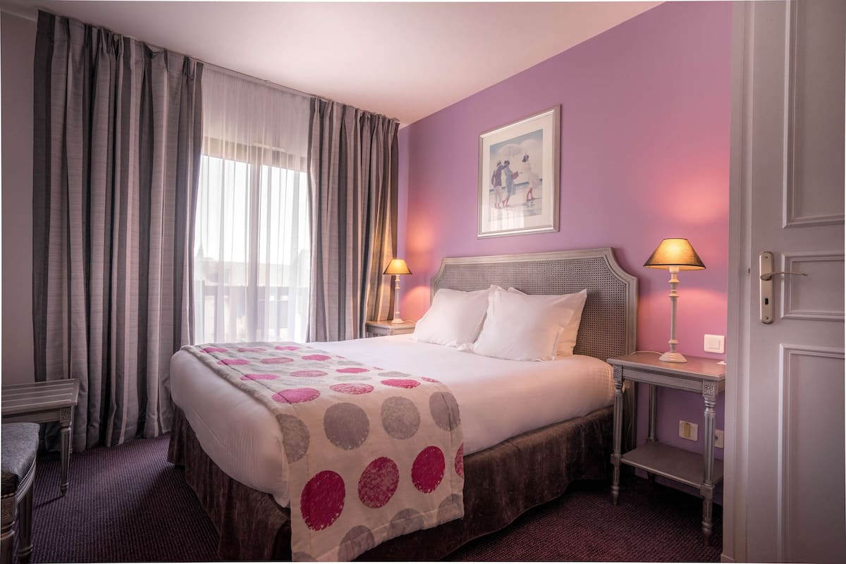Een net opgemaakt bed met een stippenkleed, een nachtkastje met een lamp en een ingelijste foto aan de muur.