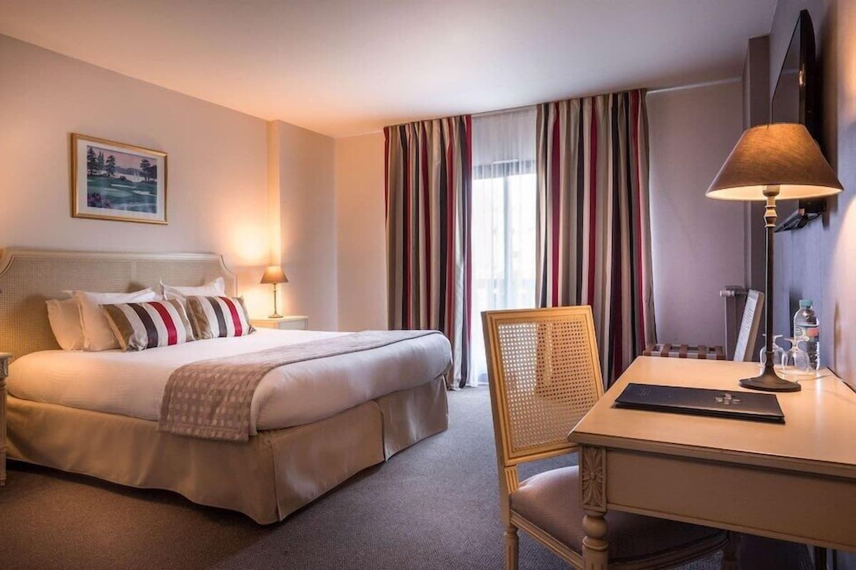 Een hotelkamer met een bed, nachtlampjes, een bureau met stoel, een raam met gordijnen en een schilderij aan de muur.