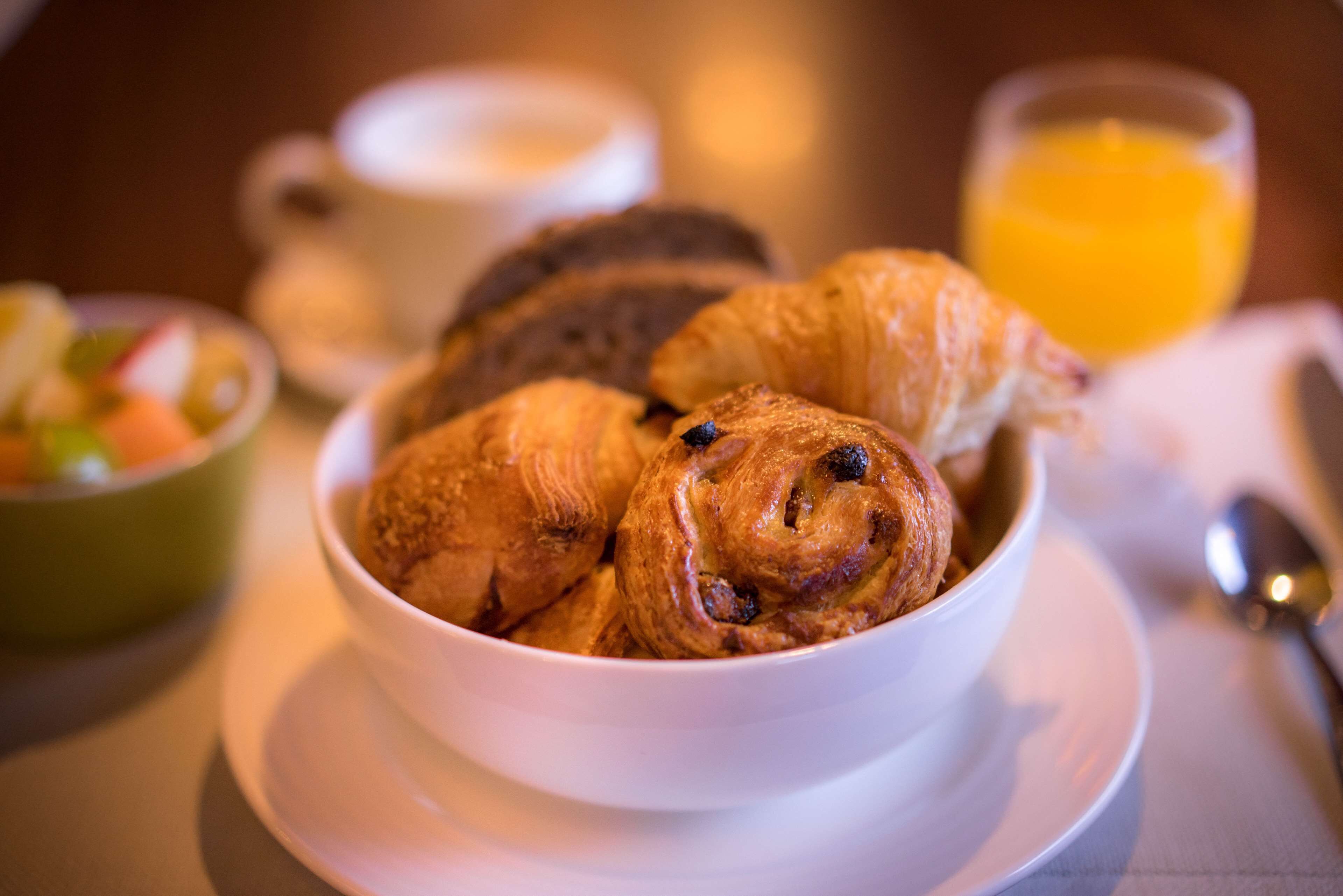 A bowl of croissants on a white plate with a cup and a glass of orange juice in the background.