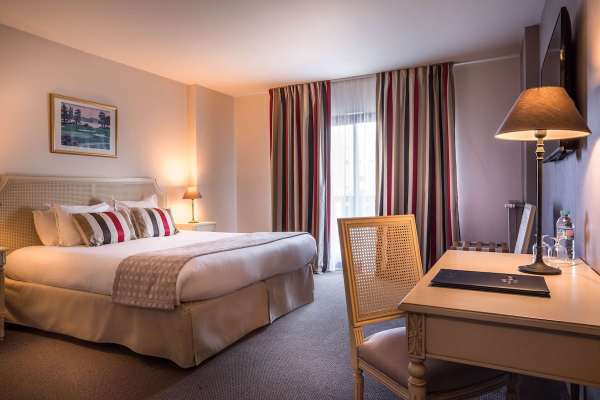 Hotelkamer met een bed, bedlampen, een bureau met een laptop, een stoel en een groot raam met gestreepte gordijnen.