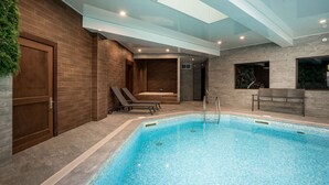 A modern indoor swimming pool with a glass ceiling, lounge chairs, and a bench.