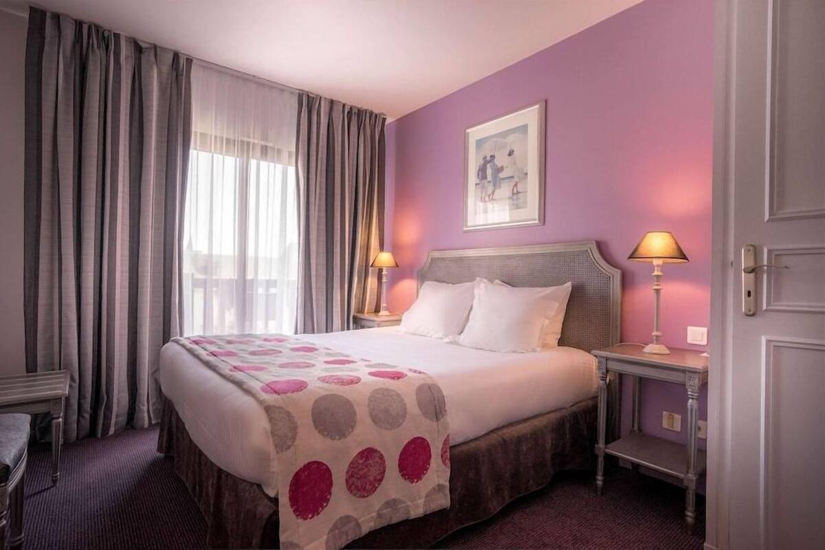 Een hotelkamer met een bed, twee bedlampen, een raam met gordijnen, een foto in een lijst aan de muur en een deur.