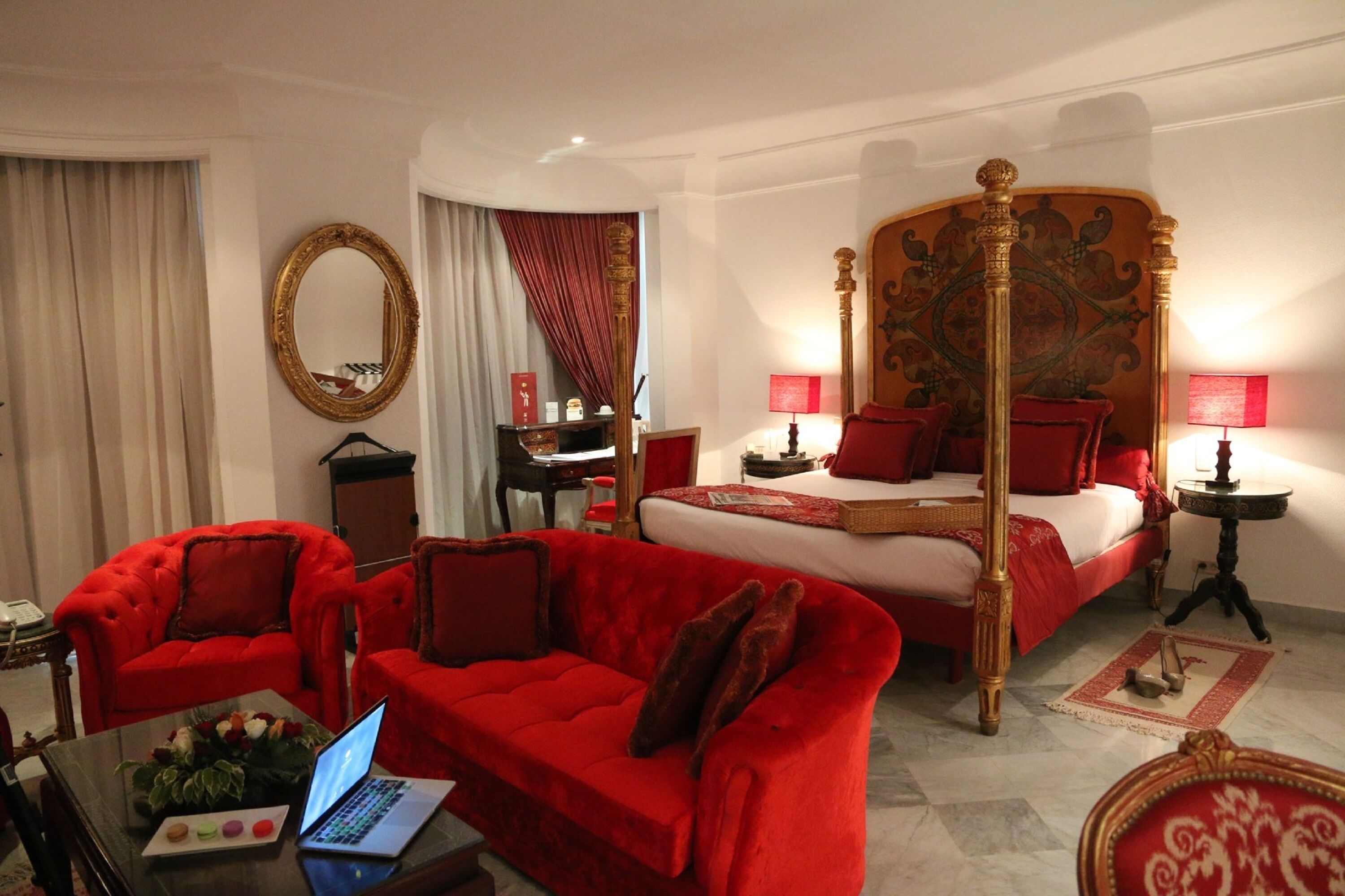 Una habitación de hotel con una cama con dosel, muebles tapizados en rojo, una computadora portátil sobre una mesa y un espejo en la pared.