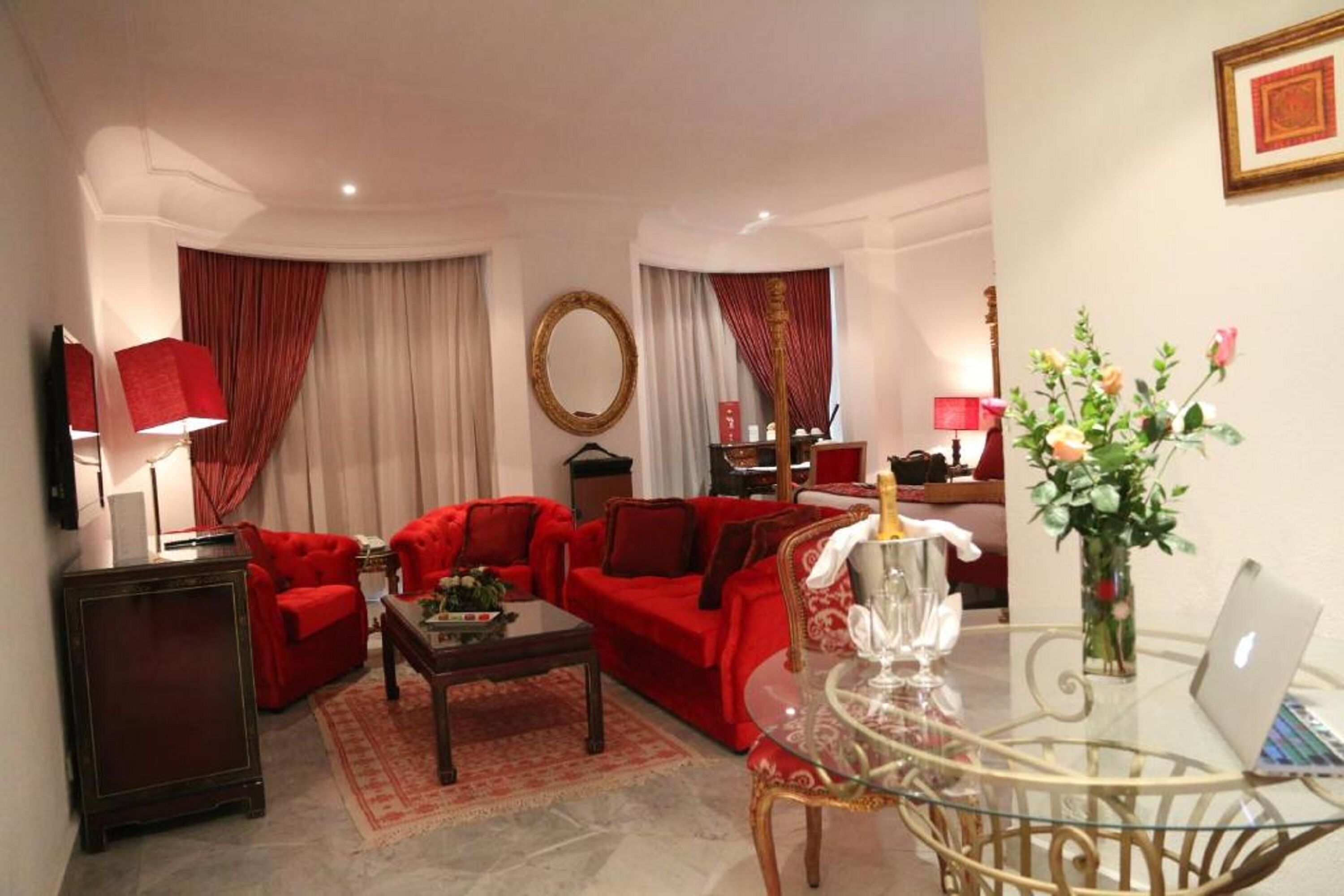 Una sala de estar con sofás rojos, una mesa de centro de vidrio, una mesa redonda con un ordenador portátil, un televisor y un espejo decorativo.