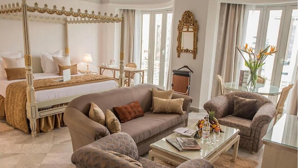 Une chambre à coucher avec un lit à baldaquin, un coin salon avec un canapé et des fauteuils, une table basse en verre, et un coin repas avec une table et des chaises.