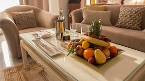 Un salon avec une table basse en verre, une bouteille, un panier de fruits et un canapé agrémenté de coussins à motifs.