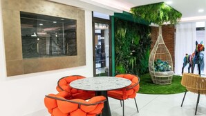 A modern interior with a round table, orange cushioned chairs, a hanging chair, and a green wall with plants. - Hotel Amazonia (CAYENNE)