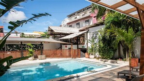 A swimming pool with lounge chairs, surrounded by tropical plants and a covered patio area. - Hotel Amazonia (CAYENNE)