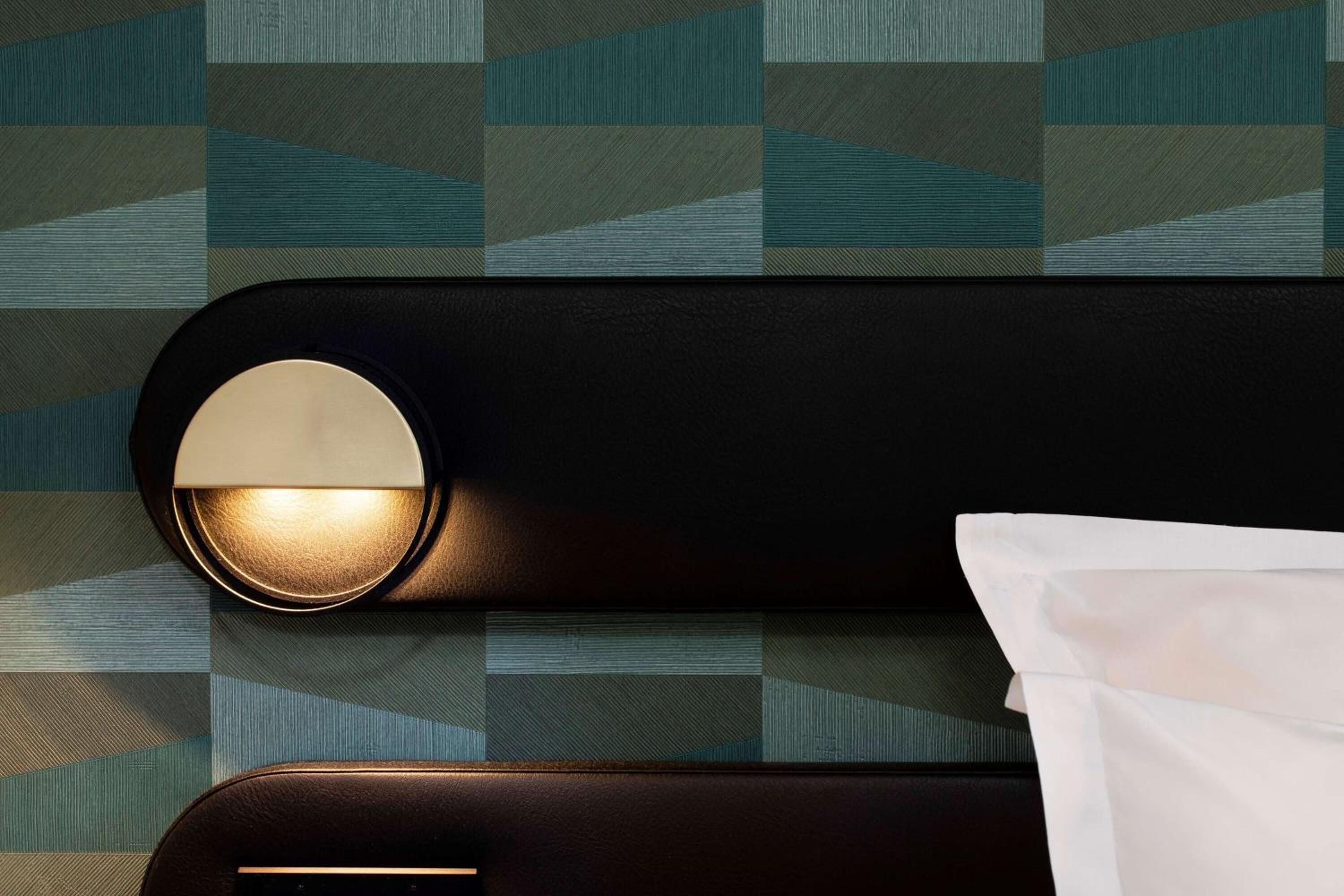 Et moderne værelse med et geometrisk mønstret væg, en sort sengegavl med en rund lampe, og en hvid pude.