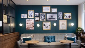A cozy seating area with a sofa and two round tables, a wall adorned with framed pictures and posters, and a wooden partition on the left. - Maison 46 (Paris)