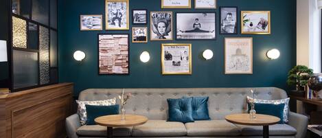 A cozy seating area with a sofa and two round tables, a wall adorned with framed pictures and posters, and a wooden partition on the left.
