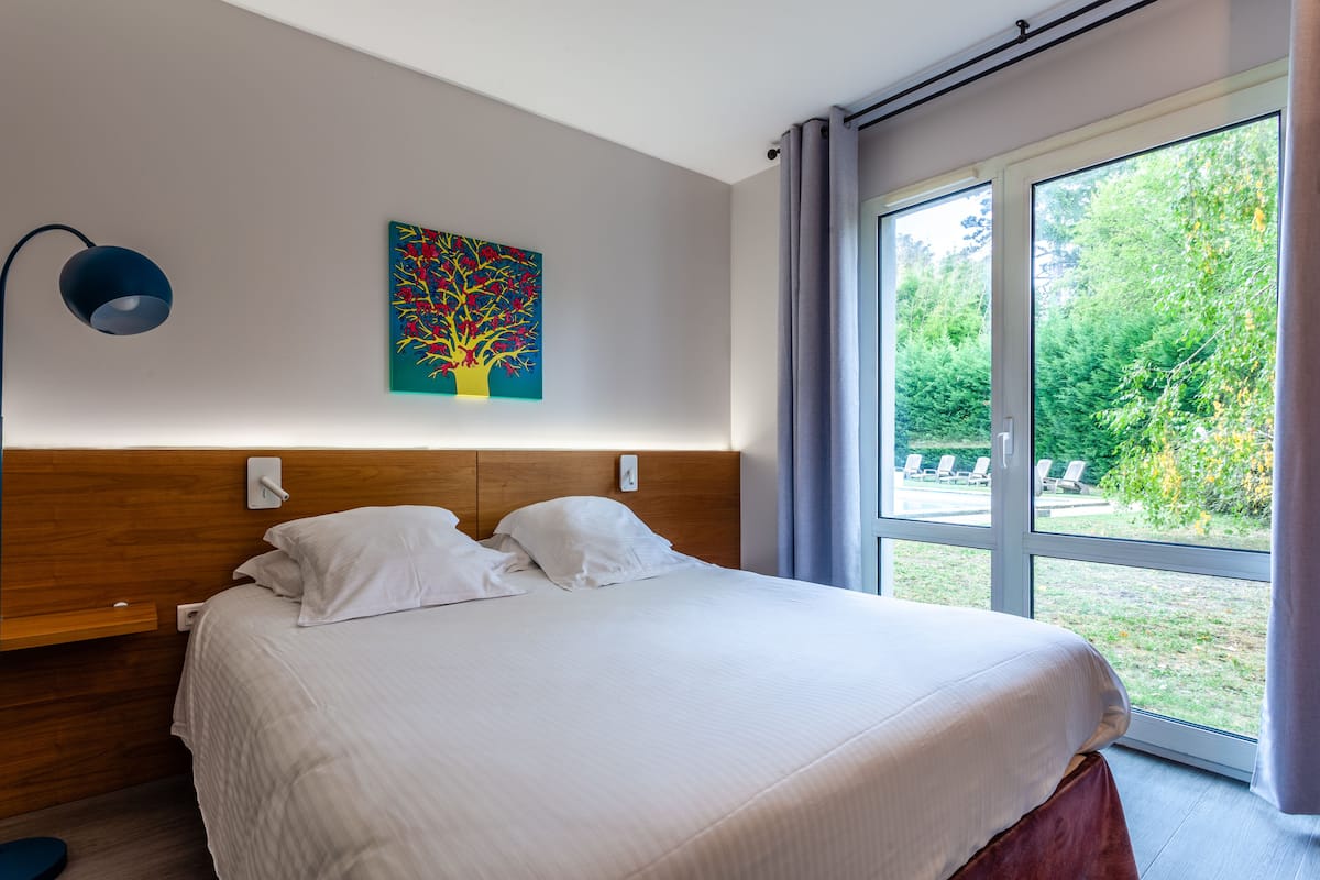 A modern bedroom with a large bed, a nightstand, a wall-mounted lamp, and a window with a view of the outdoors.
