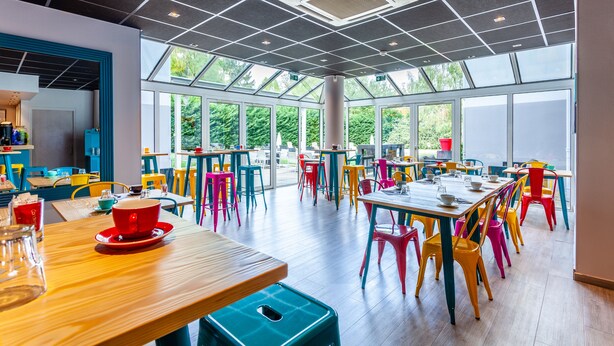 Un vaste espace intérieur aménagé en salle à manger, avec de hauts plafonds, de grandes fenêtres, ainsi que des chaises et des tables aux couleurs vives.