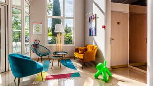 Un hall d’entrée moderne avec de grandes fenêtres, un fauteuil bleu, un fauteuil jaune, une sculpture verte et une porte en bois.