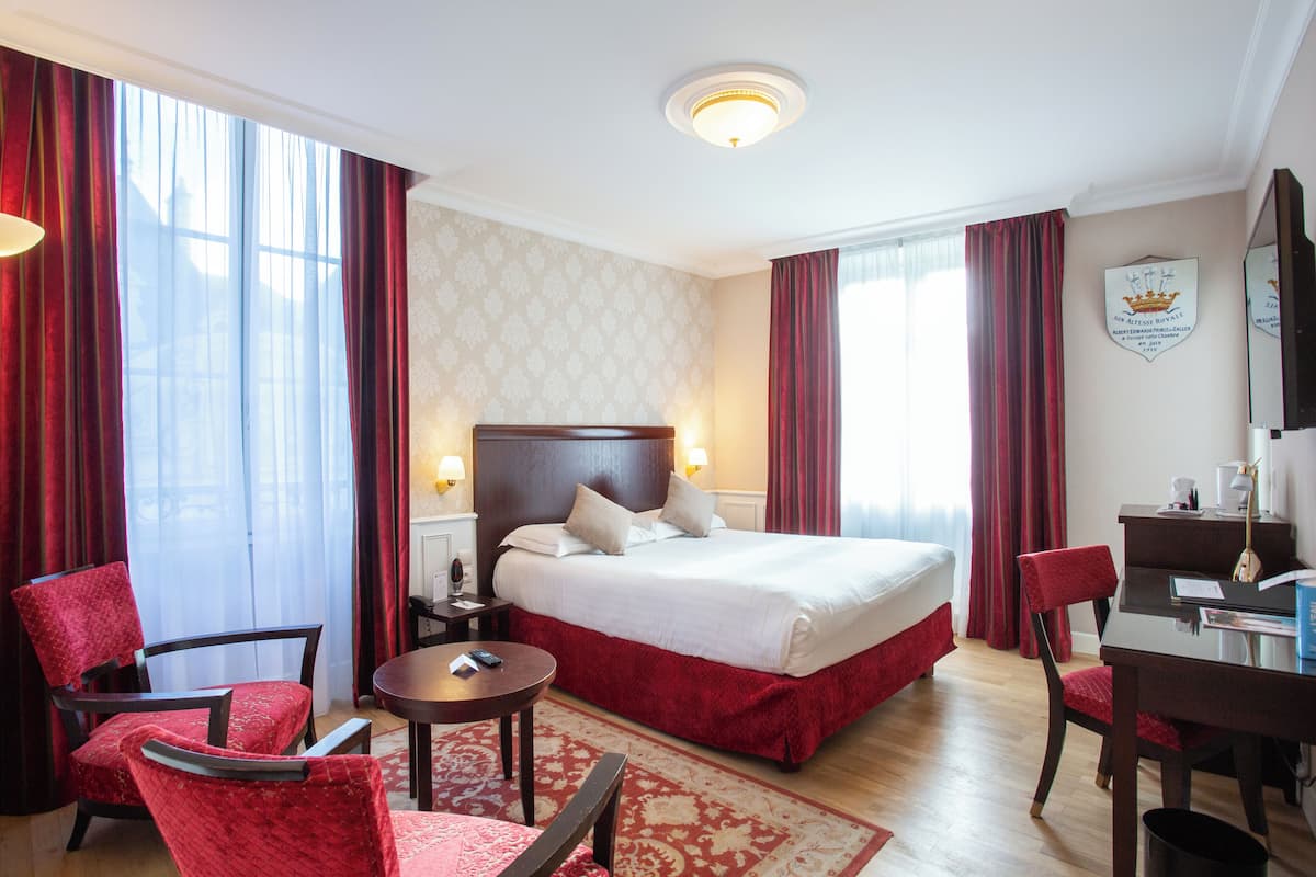 A hotel room with a large bed, two red upholstered chairs, a small round table, and a desk with a chair.