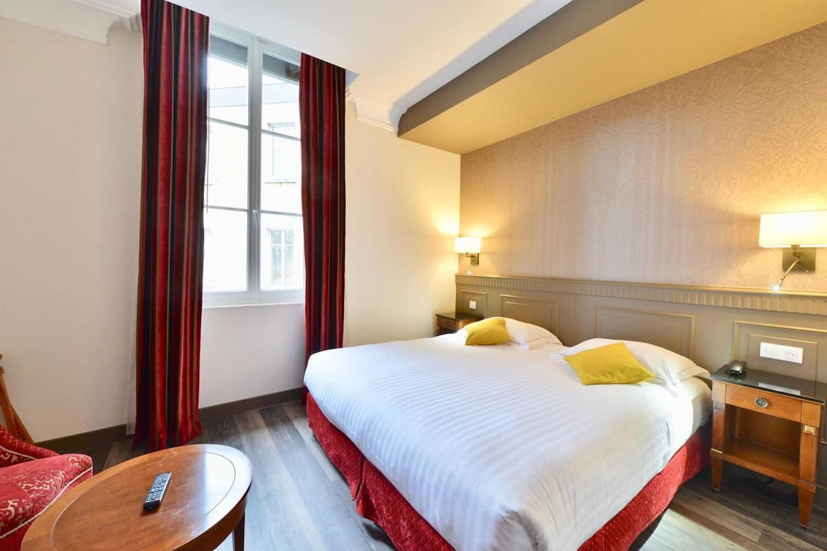 A hotel room with a large bed, two bedside tables, a nightstand, a chair, a small table, and a window with curtains.