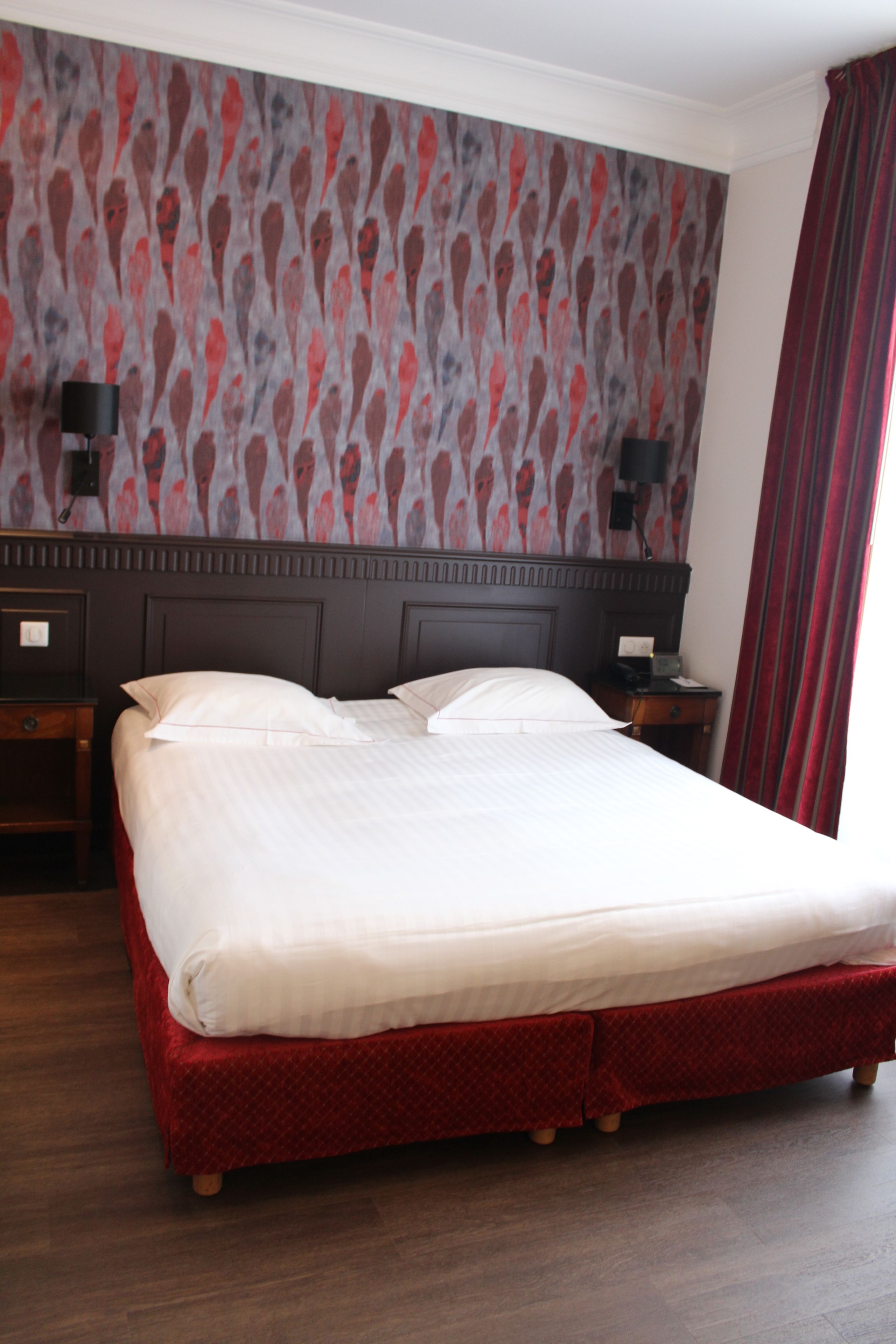 Une chambre d’hôtel avec un grand lit, une tête de lit recouverte d’un papier peint à motifs, deux tables de chevet avec des lampes et un rideau rouge.