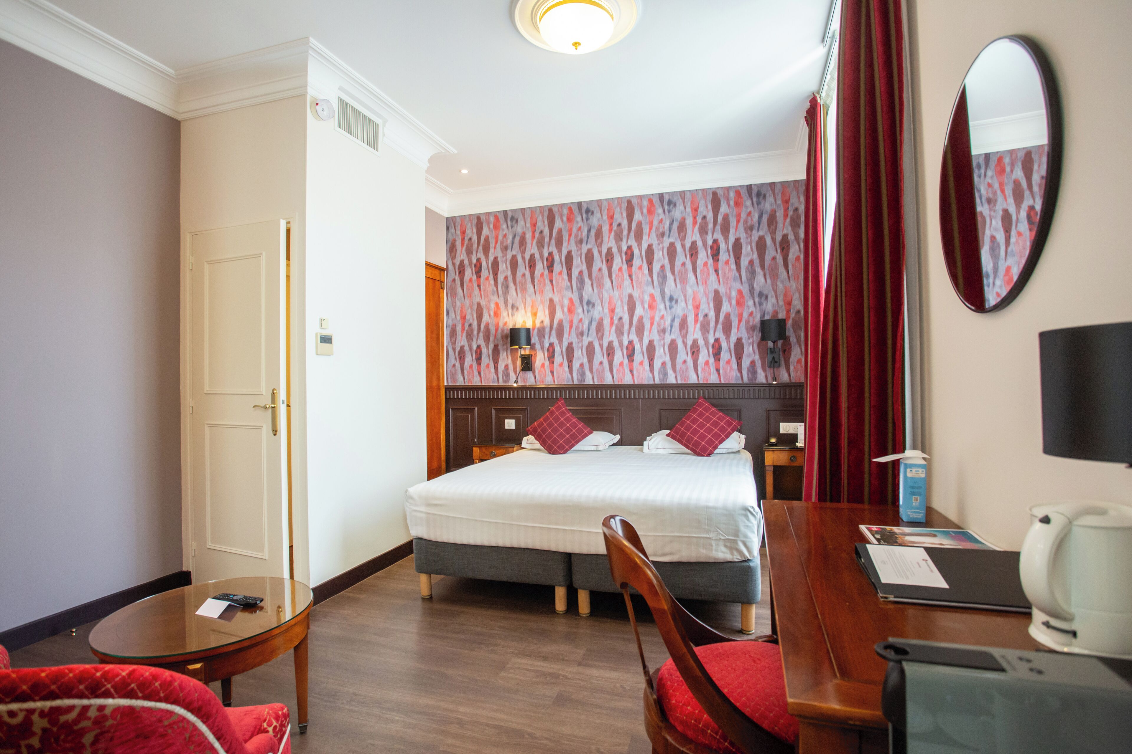 A hotel room with a bed, a desk with a chair, a small table, a mirror, and a wall with patterned wallpaper.
