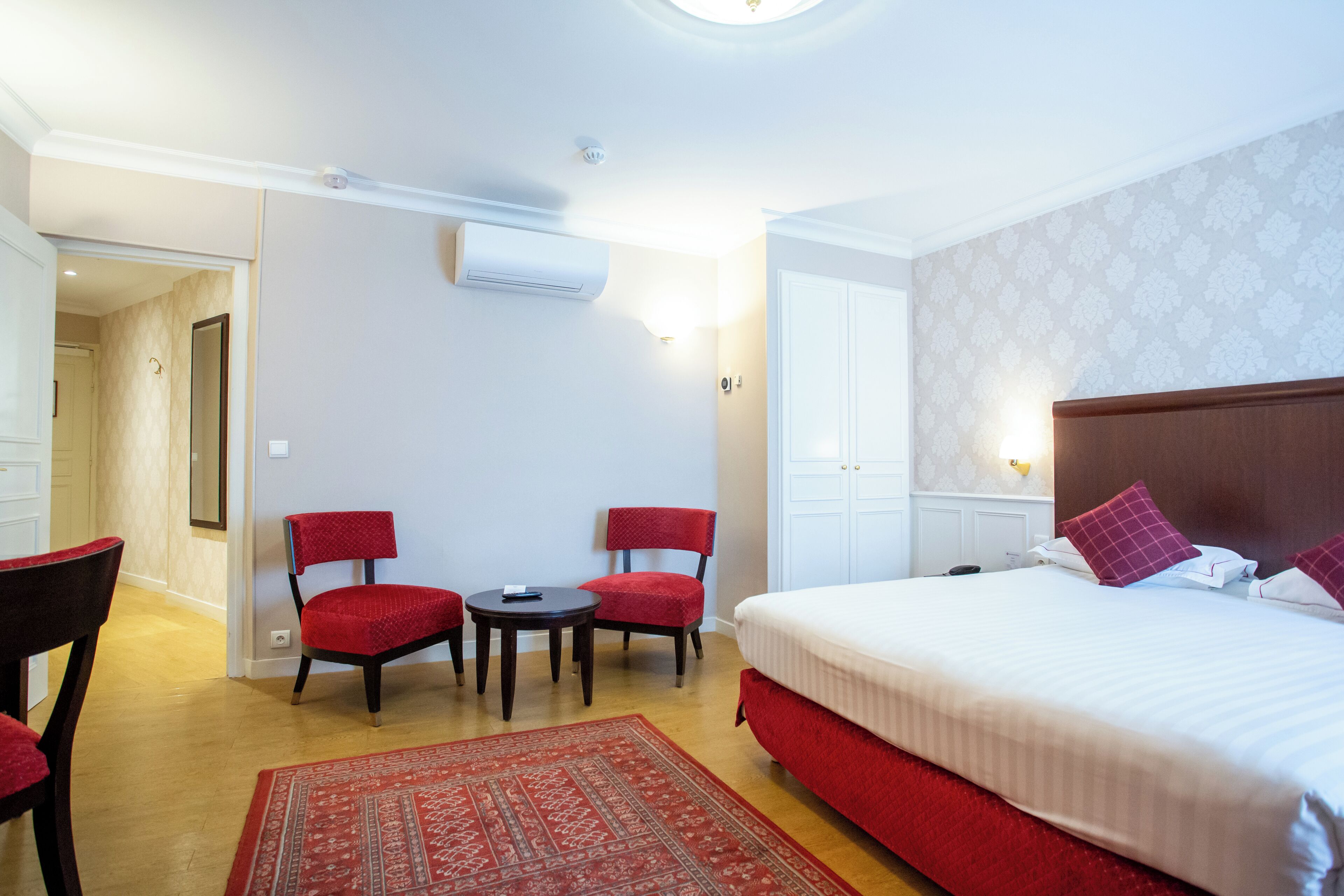 A hotel room with a large bed, two red chairs, a small table, and a red rug.