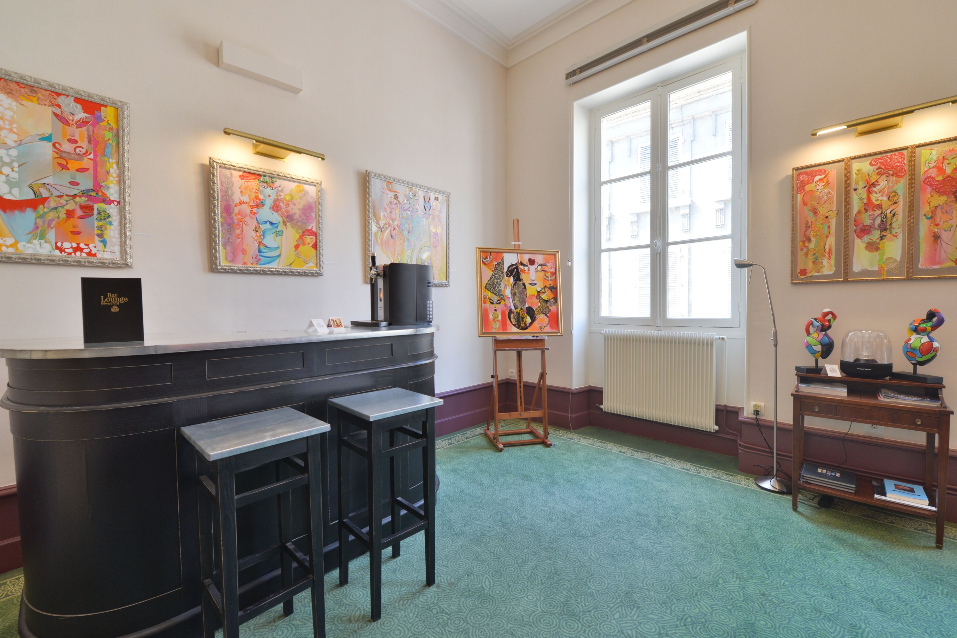 A room with a bar area, artwork on the walls, and a large window providing natural light.