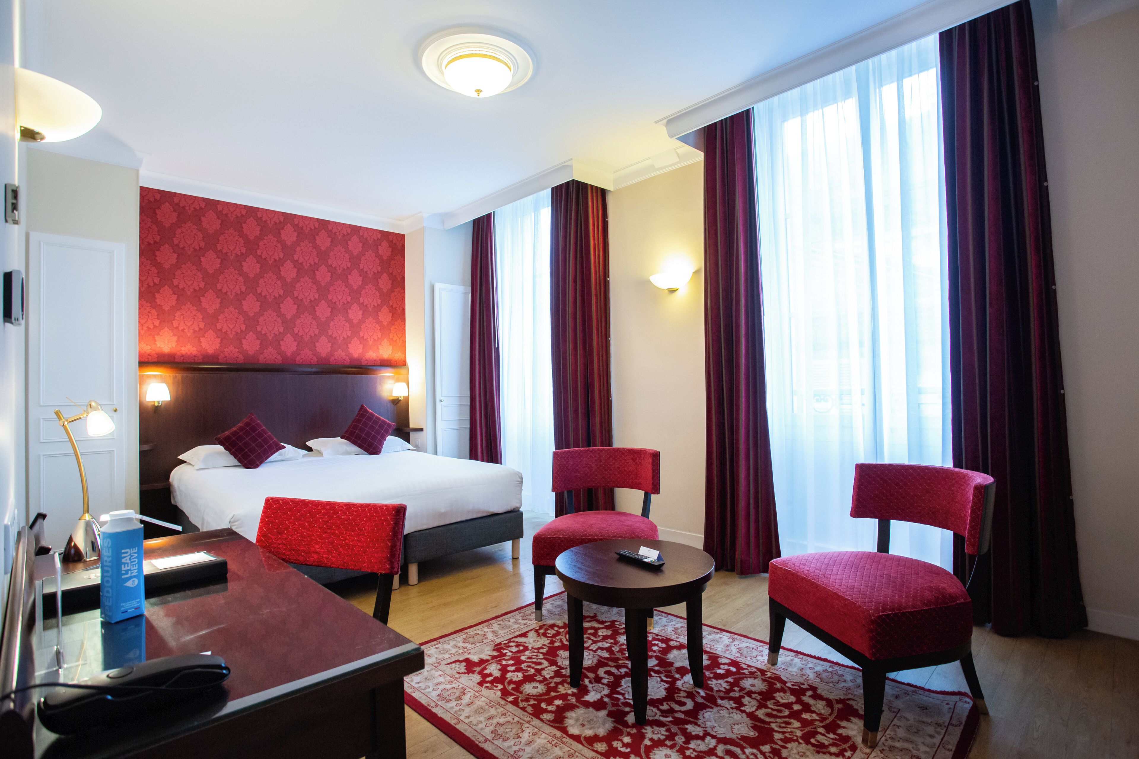 Habitación de hotel con cama, dos sillas rojas, una mesita, un escritorio con lámpara y un ventanal con cortinas.
