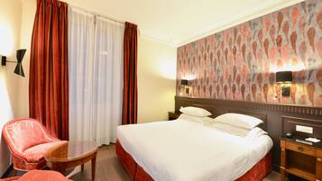 Una habitación de hotel con una cama grande, una silla roja, una mesita pequeña y una mesita de noche con una lámpara.