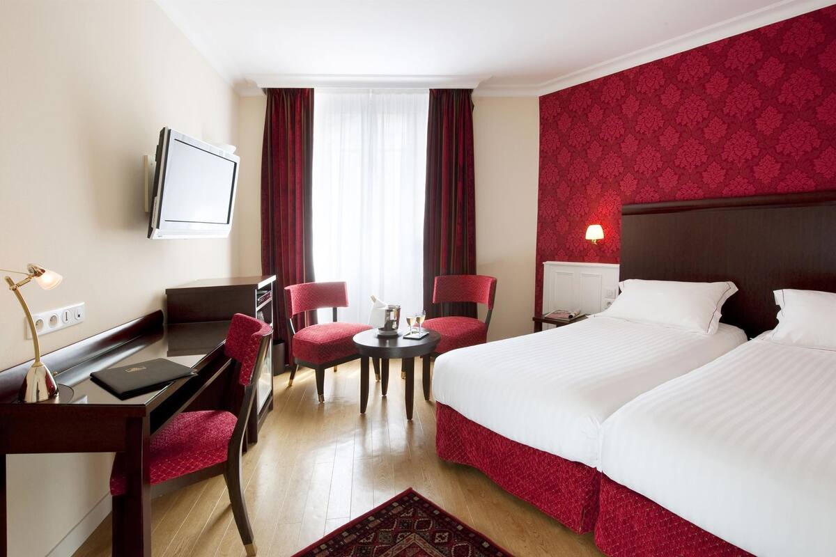 A hotel room with a bed, two chairs, a desk, a TV, and a window with curtains.