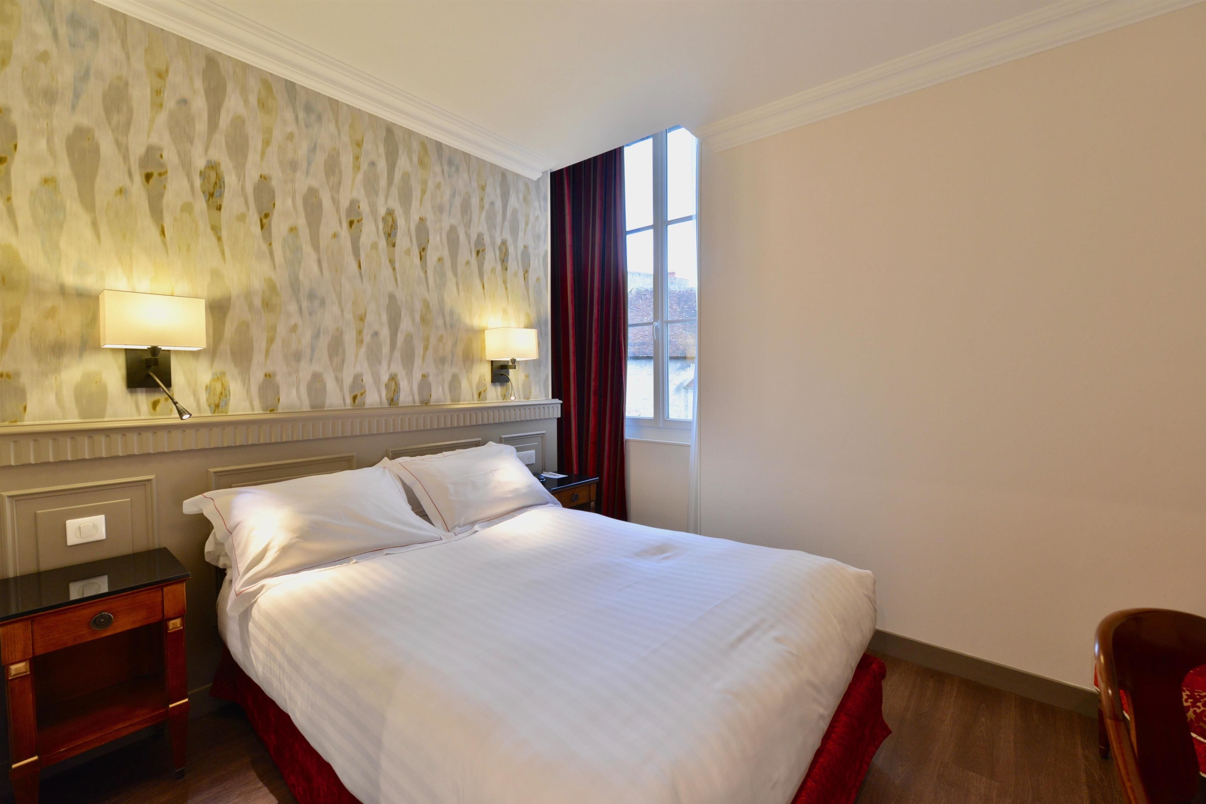 A neatly made bed with white linens, a nightstand with a lamp, a mirror, and a window with red curtains.
