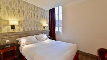 A neatly made bed with white linens, a nightstand with a lamp, a mirror, and a window with red curtains.