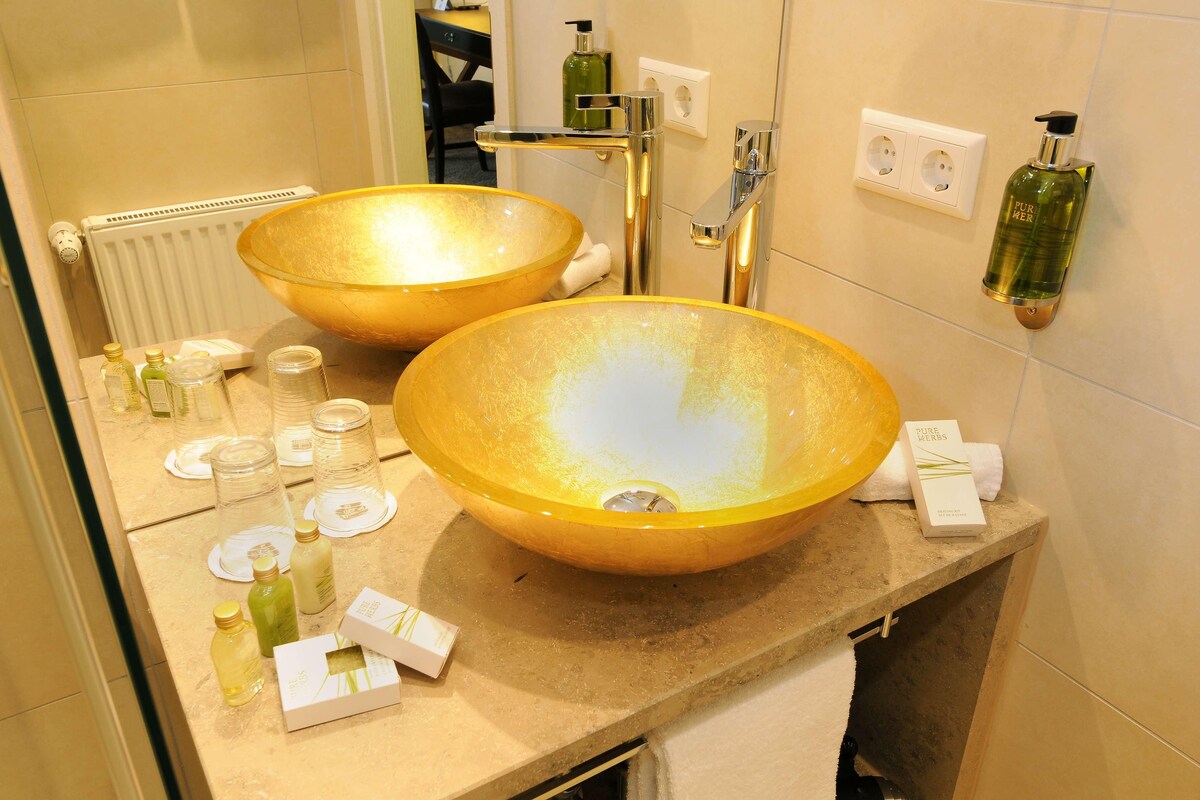 Un baño con dos lavamanos dorados grandes, un espejo, un perchero para toallas y artículos de aseo en la encimera.