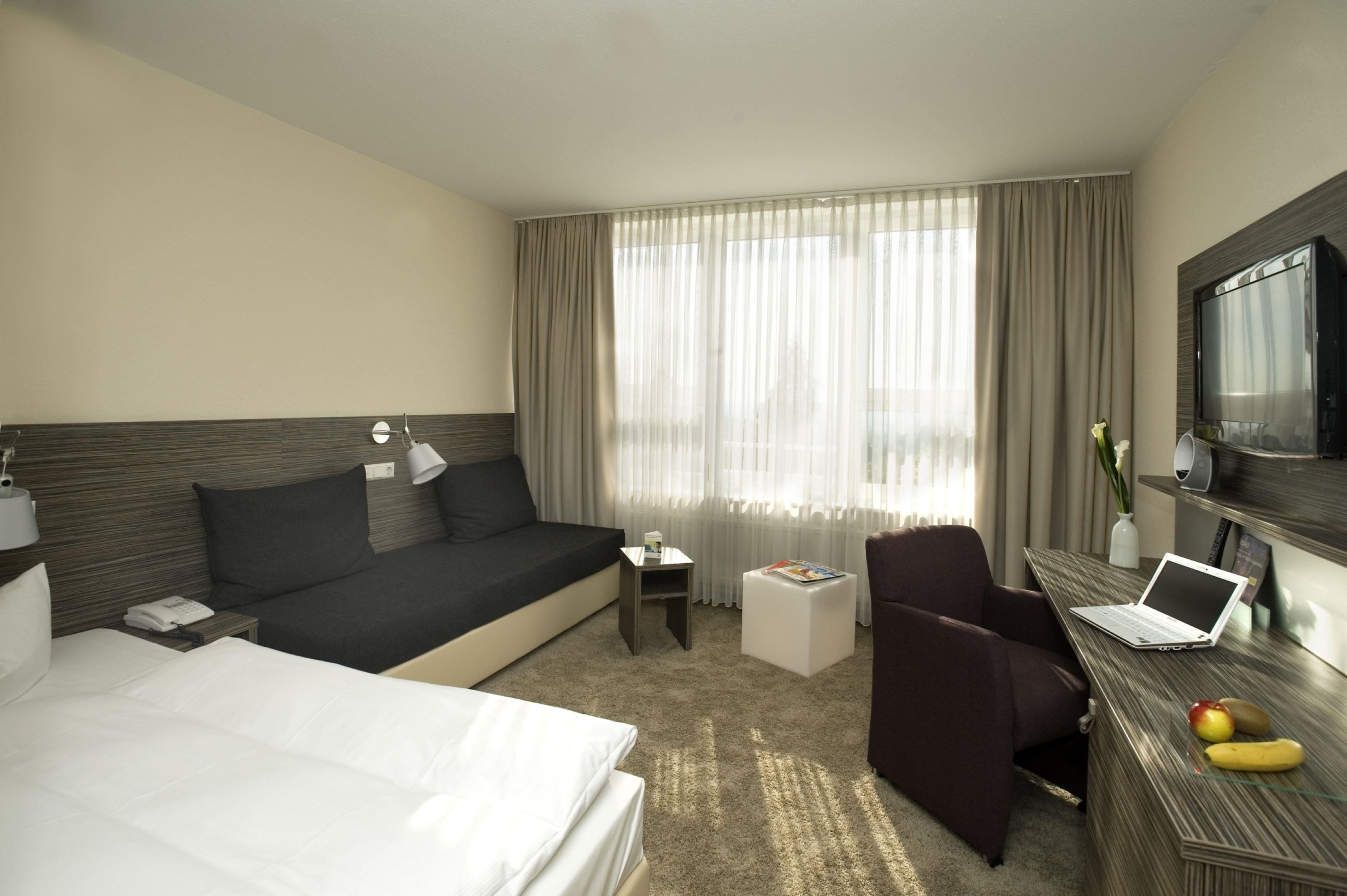 Habitación de hotel con cama, sofá, escritorio con una computadora portátil, silla, televisor y ventana con cortinas.