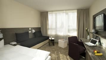 Habitación de hotel con cama, sofá, escritorio con una computadora portátil, silla, televisor y ventana con cortinas.
