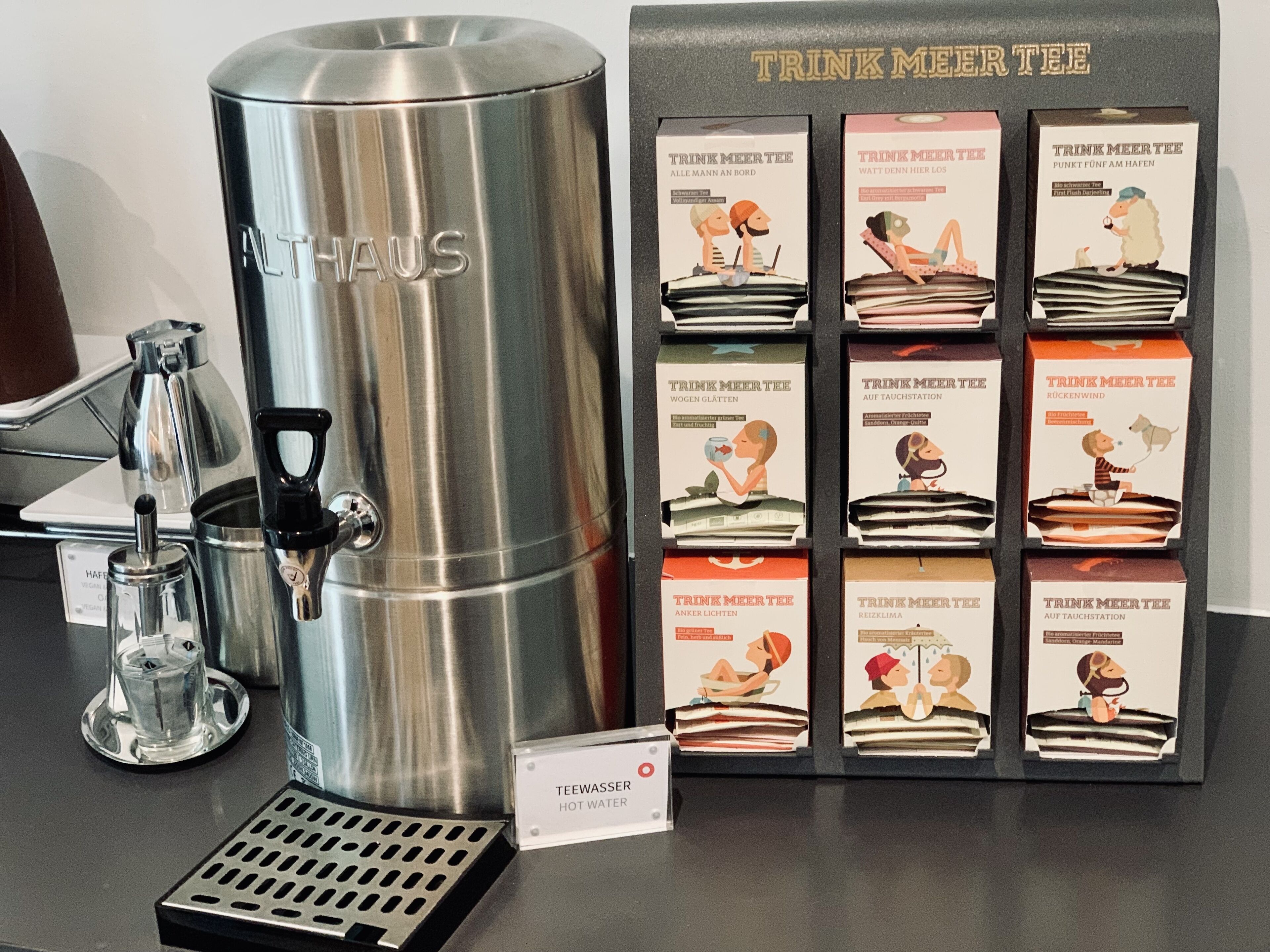 A stainless steel beverage dispenser labeled "ALTHAUS" and a display of "TRINK MEERTEE" boxes with illustrations of people enjoying drinks.