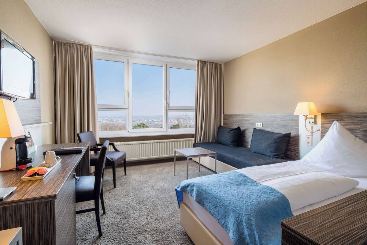A hotel room with a bed, a desk, a chair, a small table, and a view of the city through large windows.