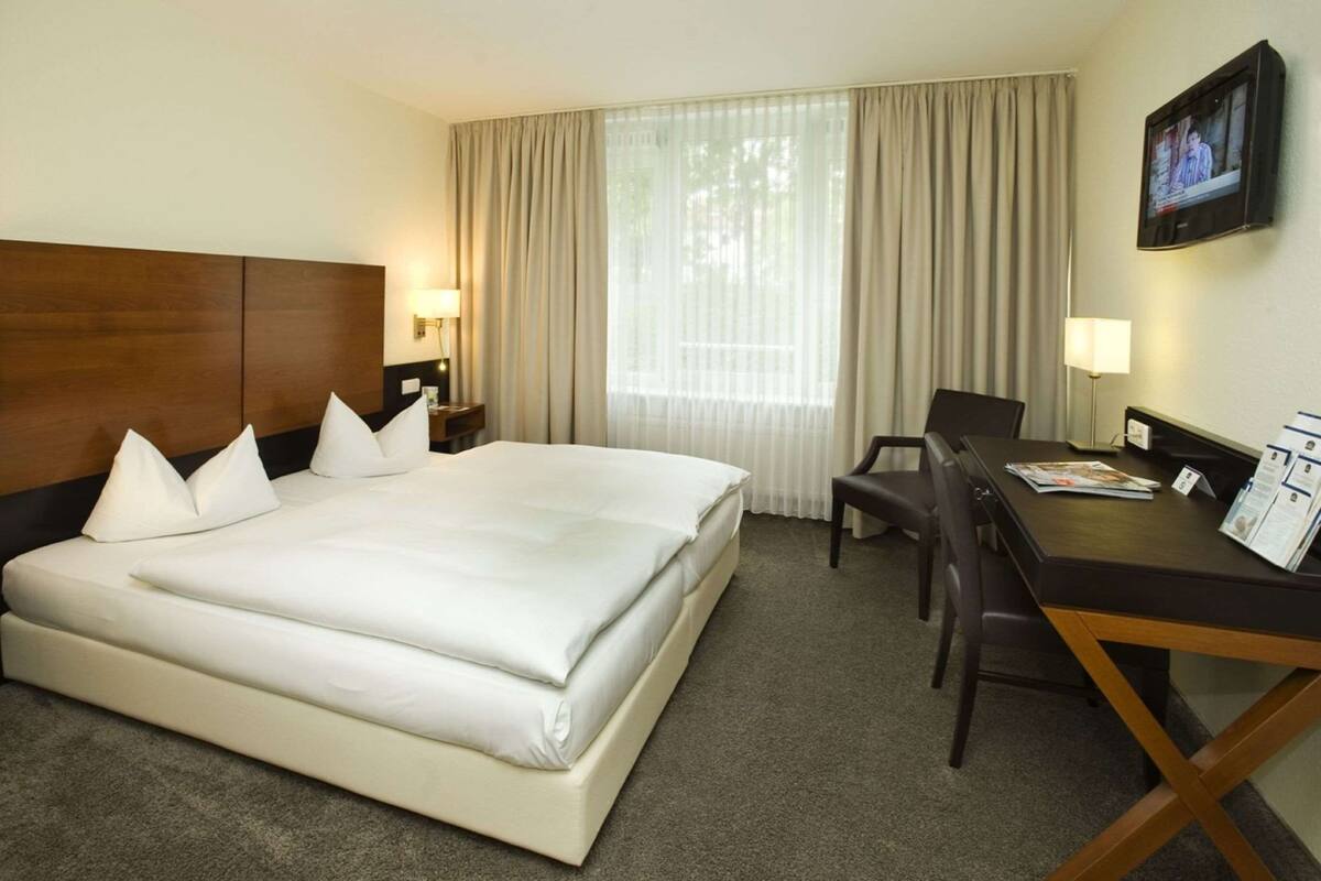 A hotel room with a large bed, a desk with a chair, a TV mounted on the wall, and a window with curtains.