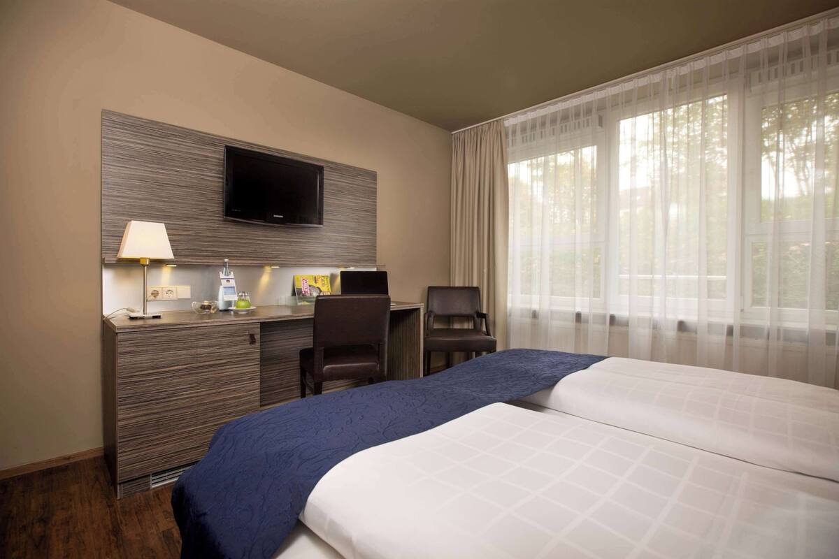 A hotel room with a bed, a desk with a lamp, a flat-screen TV mounted on the wall, and a window with sheer curtains.