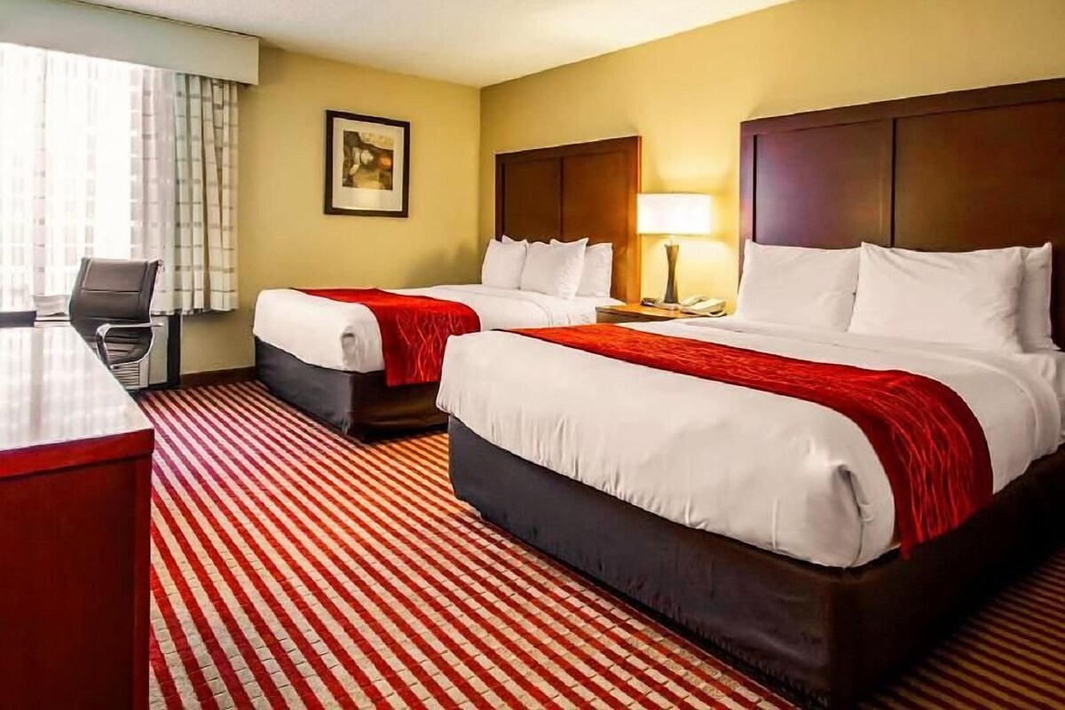 Quarto de hotel com duas camas, uma escrivaninha, uma luminária e um quadro na parede.