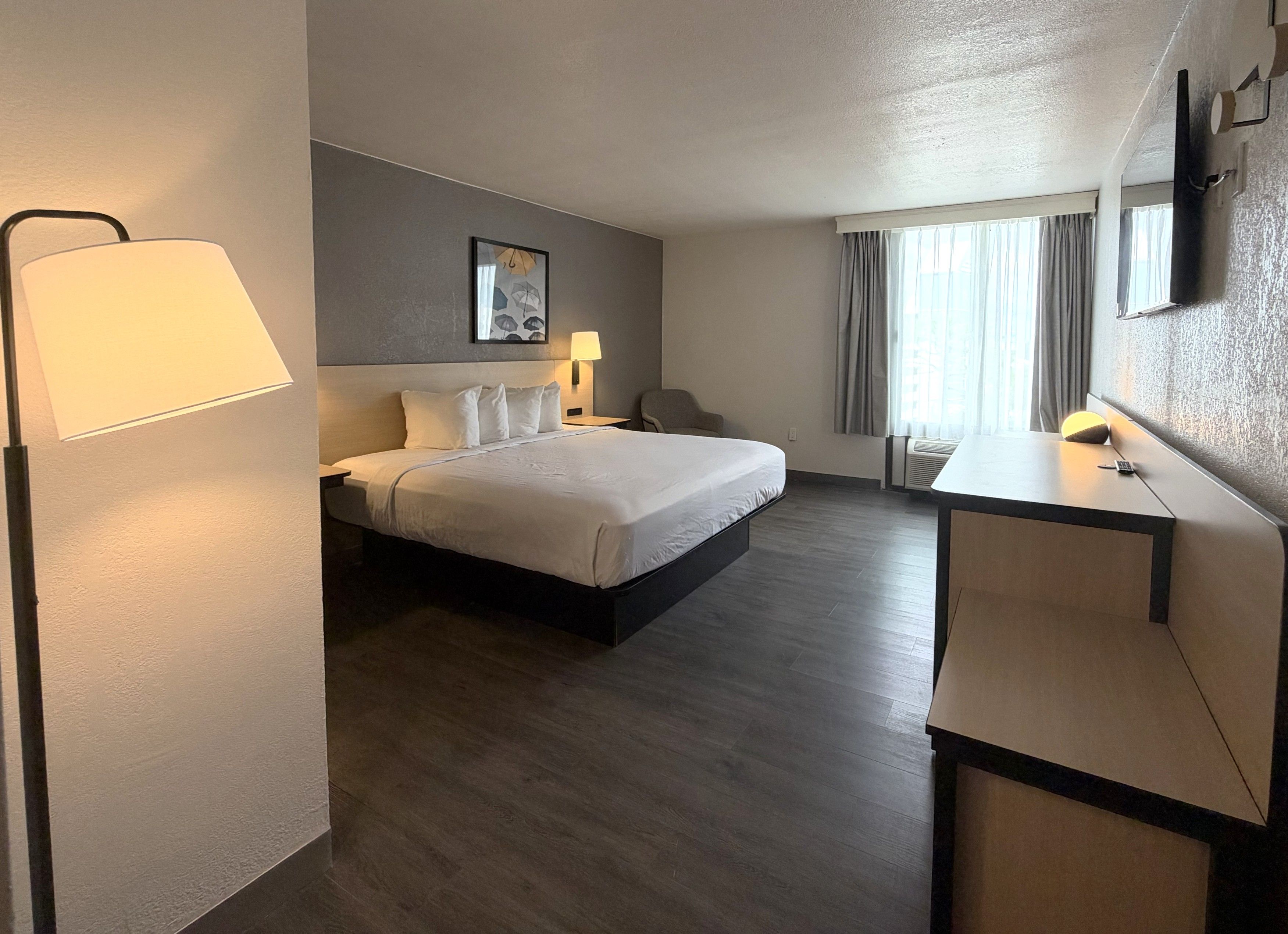 A hotel room with a bed, bedside table, lamp, and a desk with a chair.