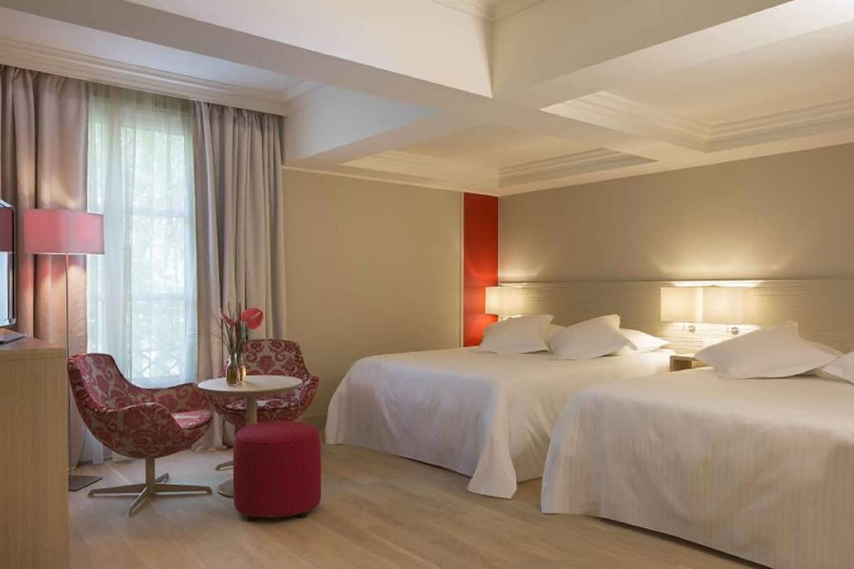 A hotel room with two beds, a desk with a lamp, a chair, and a small table with a vase of flowers.