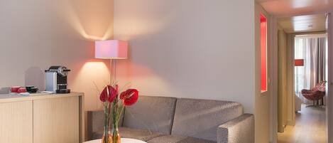 A modern hotel room with a sofa, a small round table, a lamp, and a vase with flowers.