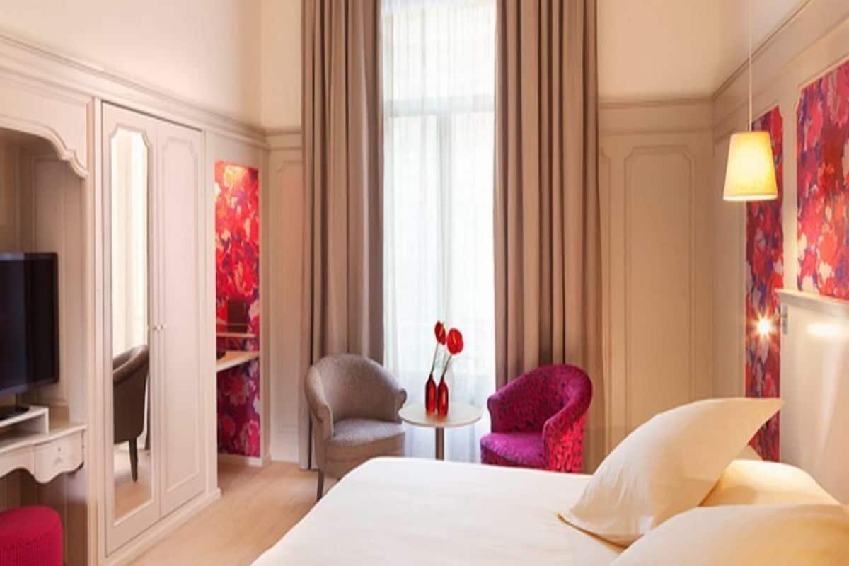 A hotel room with a bed, two armchairs, a small table with a vase, a television, and a window with curtains.