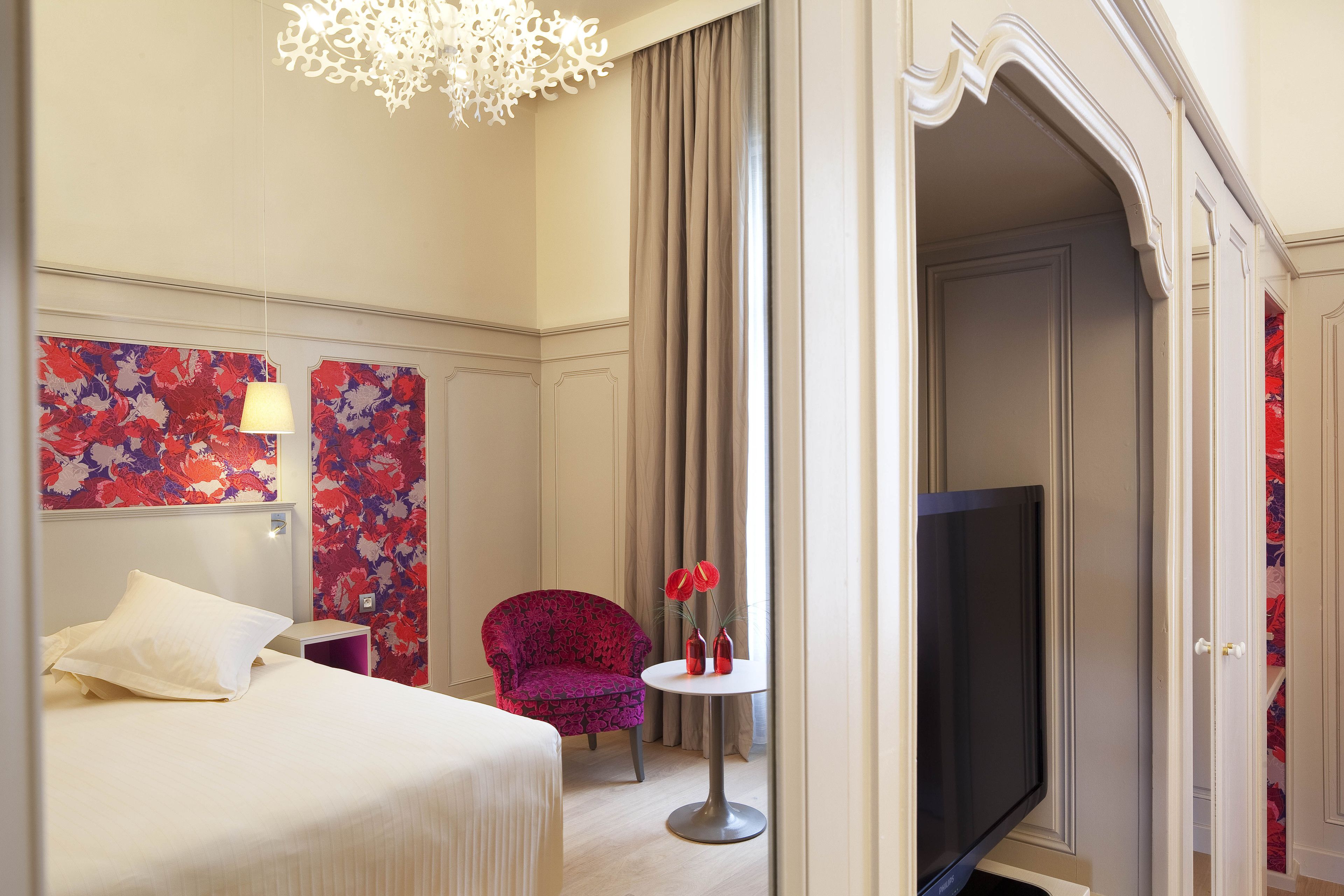 A hotel room with a bed, a chair, a small table with vases, a TV, and a decorative wall.