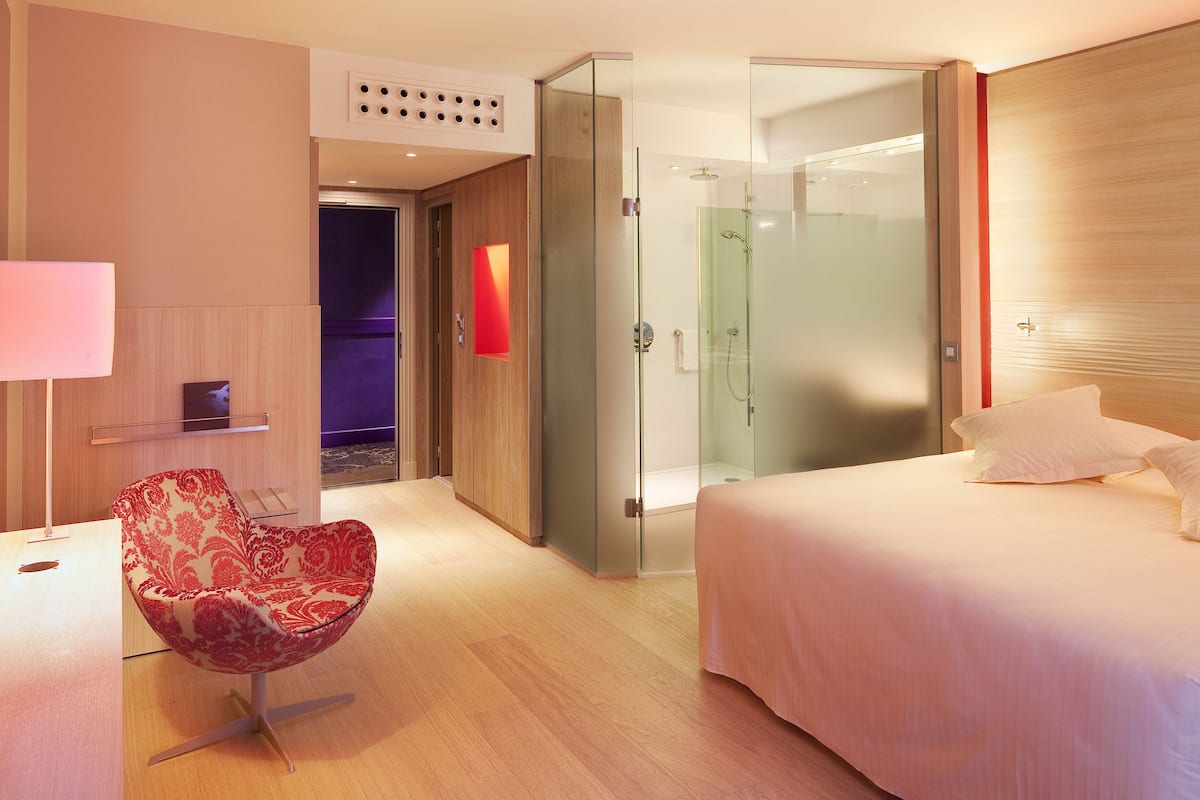 A modern hotel room with a bed, a glass-enclosed shower, a red upholstered chair, and a bedside table with a lamp.