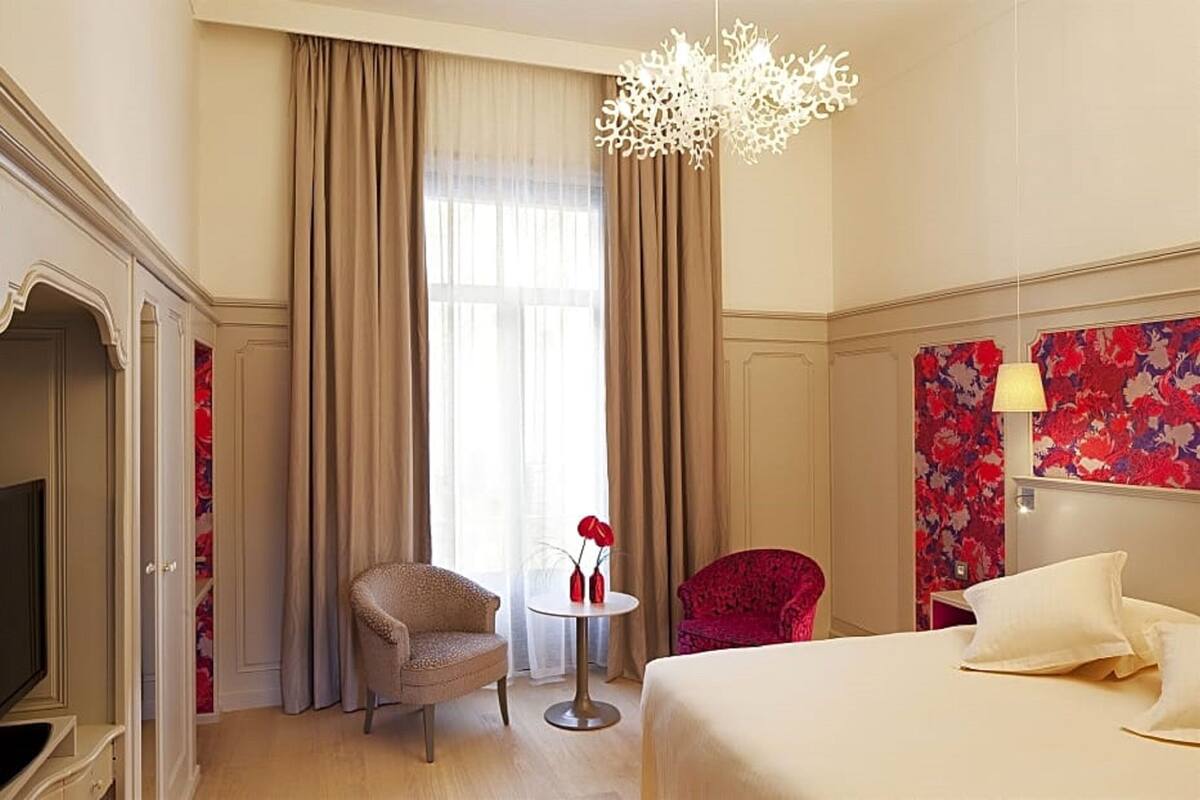 A hotel room with a bed, a red armchair, a small round table with a vase of red flowers, and a chandelier.