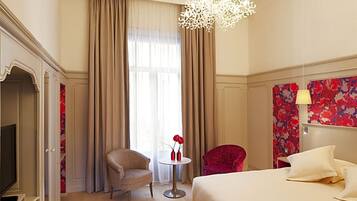 Habitación de hotel con cama, un sillón rojo, una mesita redonda con un jarrón de flores rojas y un candelabro.