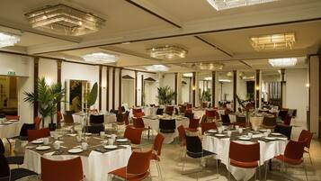 A banquet hall with multiple round tables, each set with white tablecloths, red and black chairs, and various dining utensils. The room features large mirrors on the walls and a chandelier with multiple light fixtures.